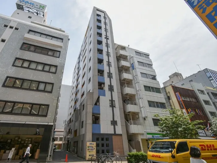- 都市型マンションの外観、周囲に立ち並ぶビル群と明るい空、近くの道路に停車中の配達車両が見える景観