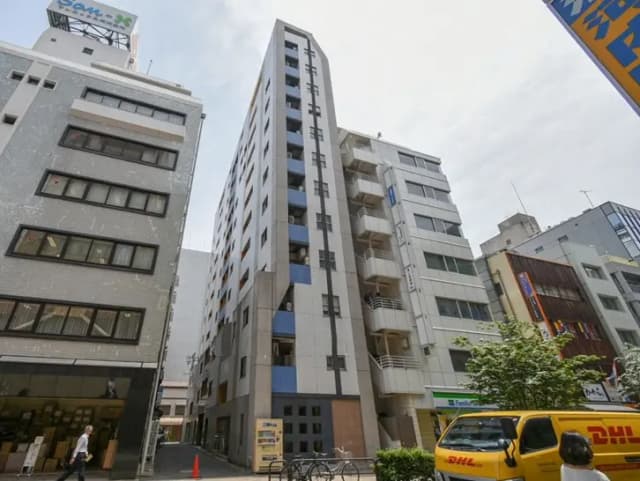- 都市型マンションの外観、周囲に立ち並ぶビル群と明るい空、近くの道路に停車中の配達車両が見える景観