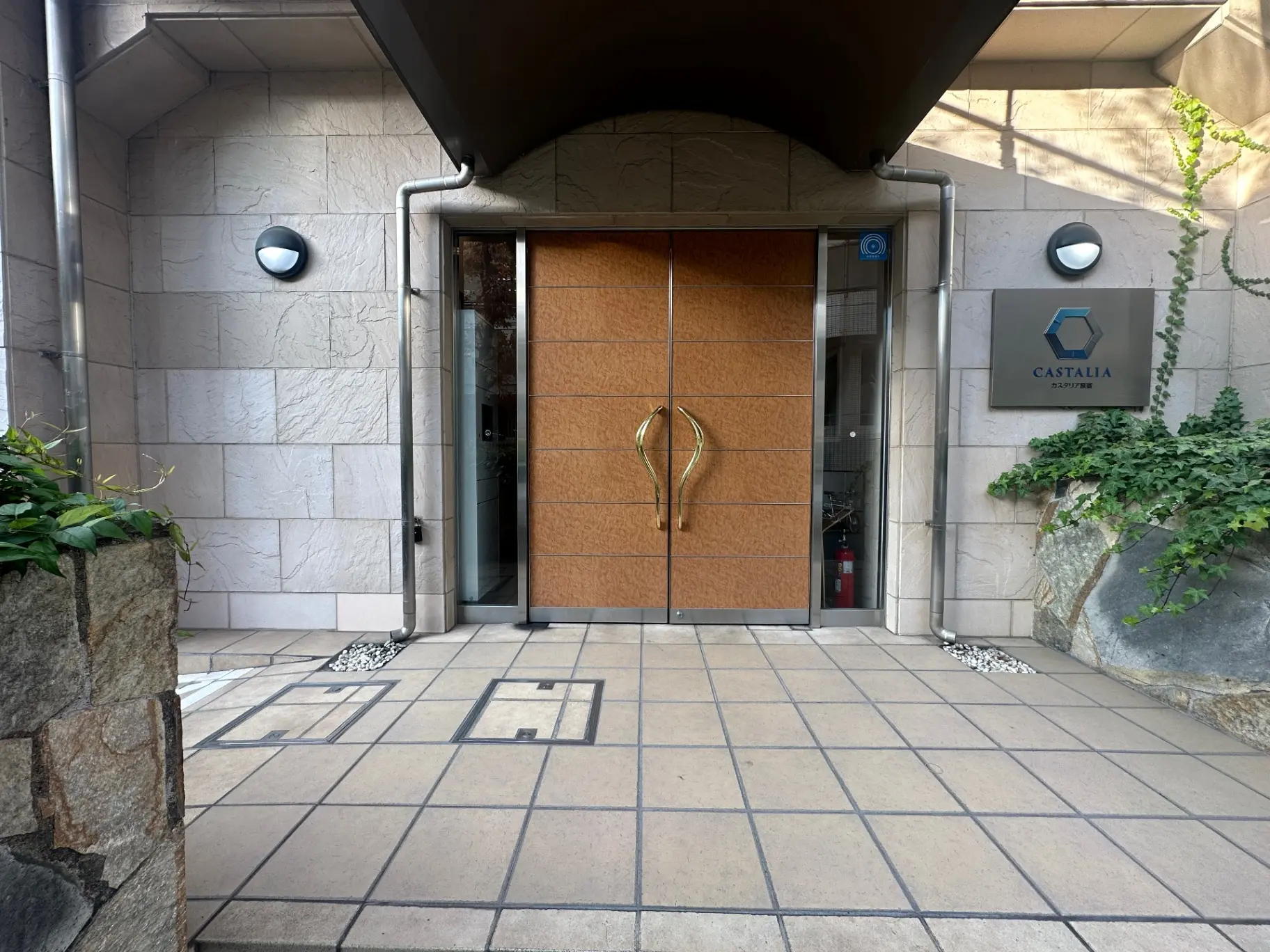 モダンなビルのエントランス、石の壁と木目調のドア、ロゴ入り看板と緑がアクセント
