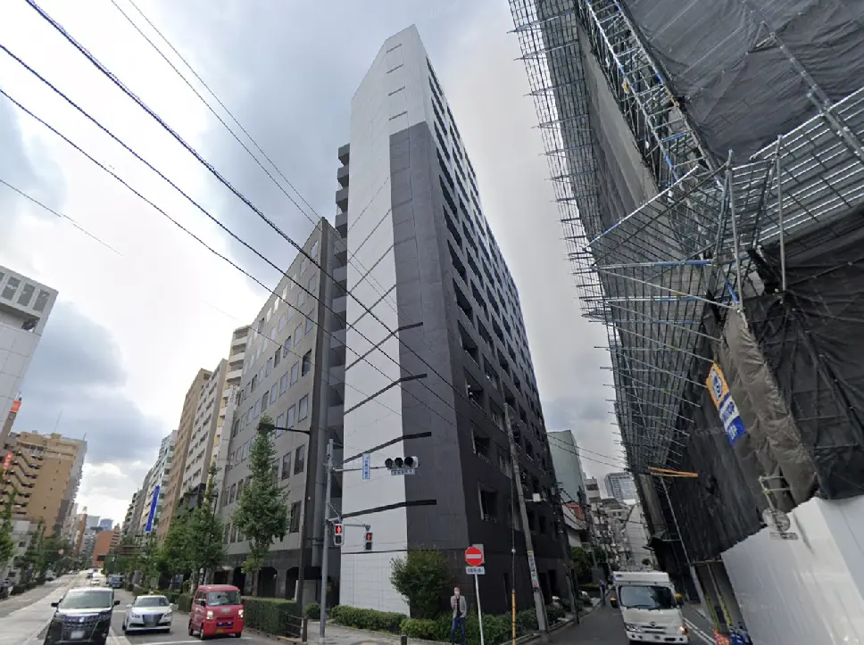 - 都市の風景に佇む現代的なビル、黒の外壁とガラスのアクセントが特徴的で、周囲には緑や交通も見える繁華街の一角