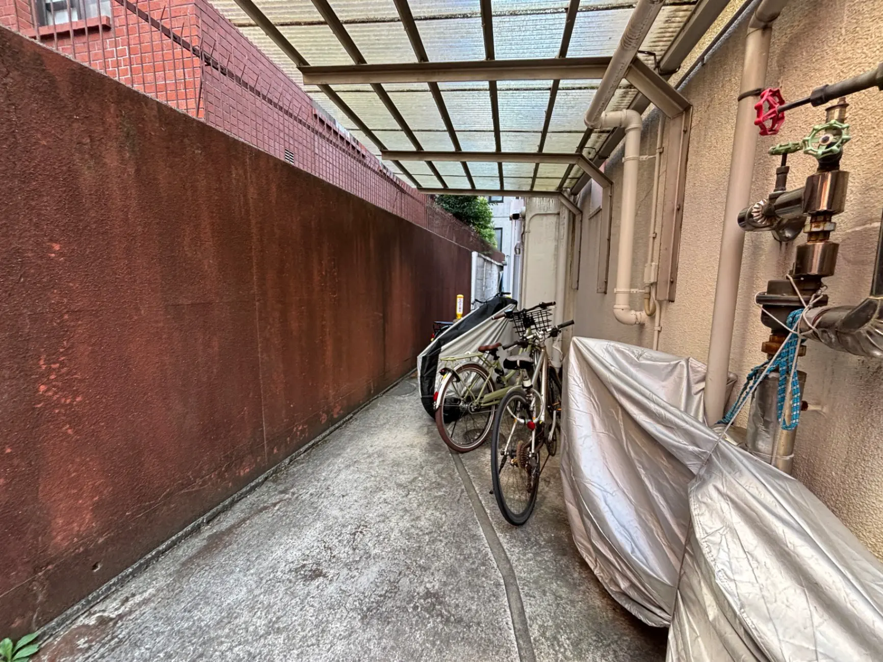 狭い通路に置かれた自転車と銀色のカバーがかかった物体、背後には赤茶色の壁と配管が見える景観