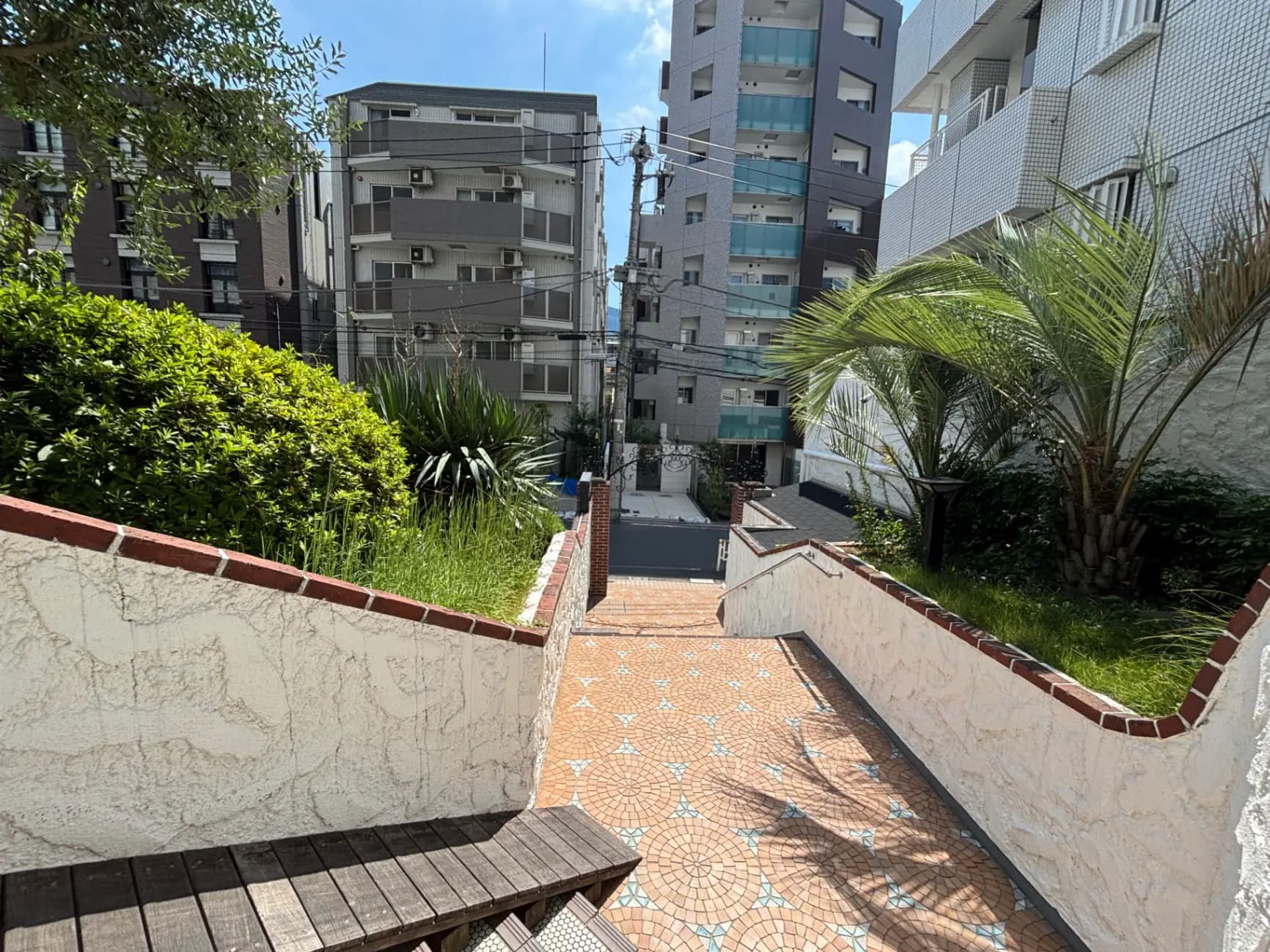 - アパートメントビルと庭の緑が見える階段、手前にはタイル敷きの歩道と周囲の建物が並ぶ風景