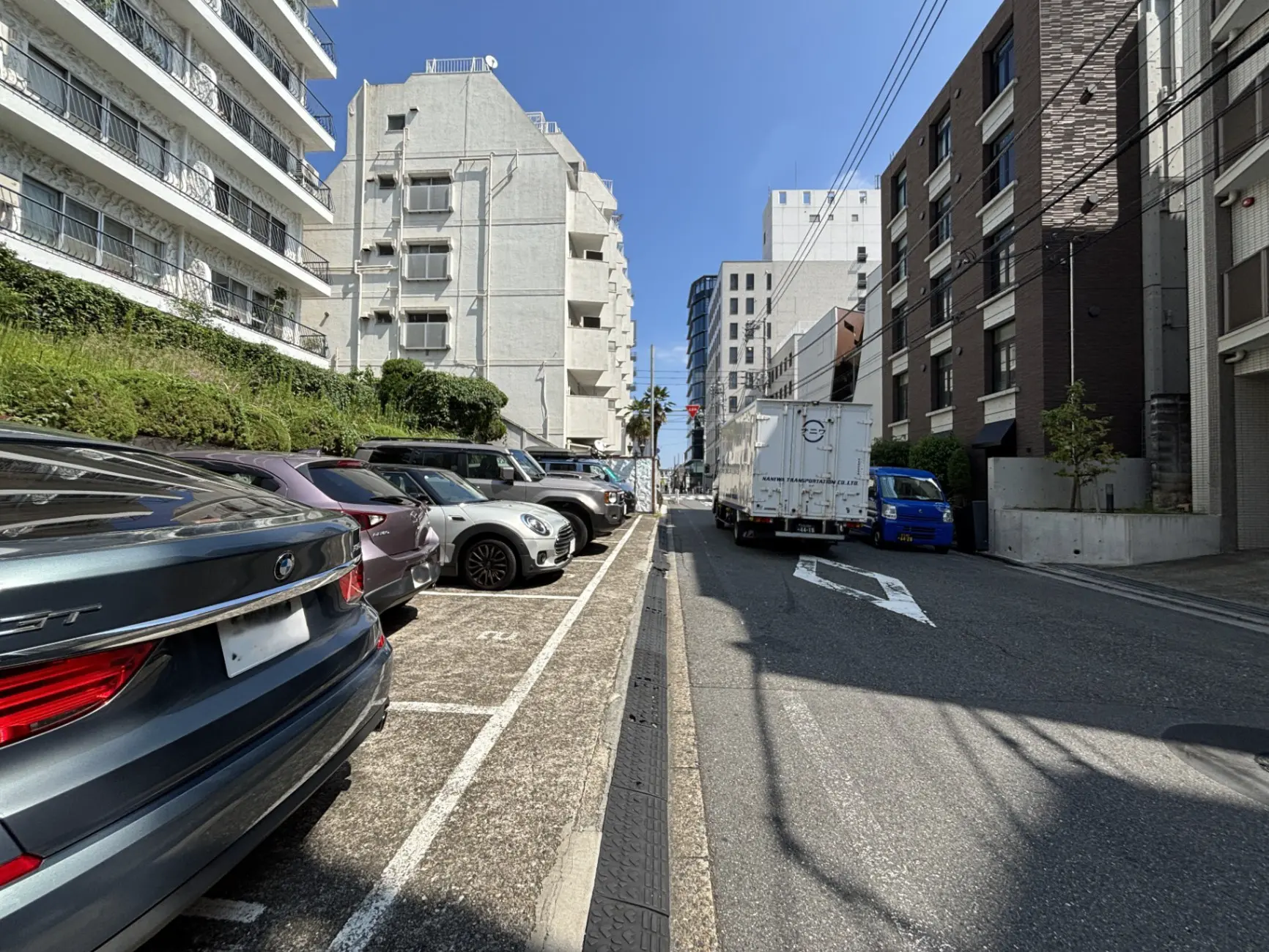 - 都会の駐車場と住宅街の風景、並んだ複数の車、奥に見えるビル群と青空のコントラスト