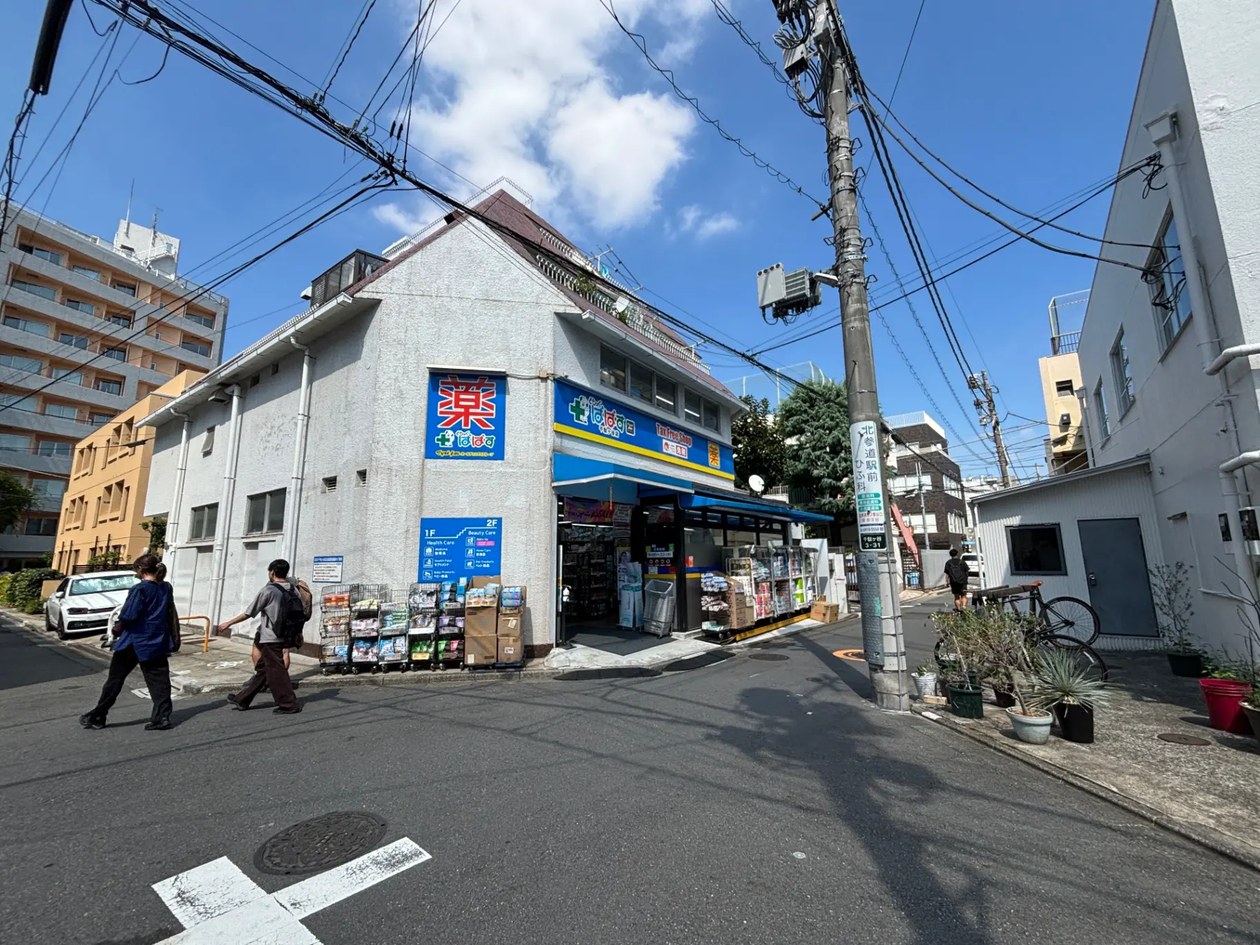 - 雑多な商品が並ぶ街角のドラッグストア、青空の下で人々が通りを行き交う風景
