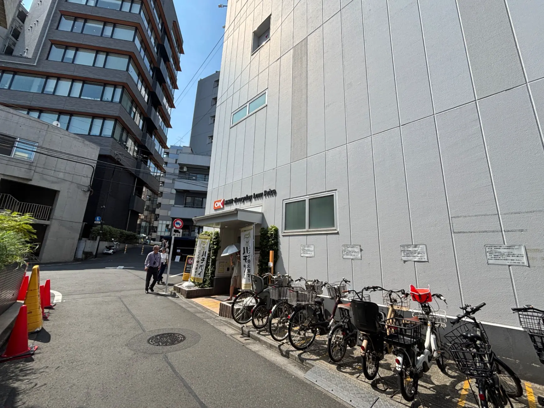 - 都市の一角にある白い建物の外観、自転車が並ぶ通りと歩行者が行き交う風景、青空を背景にした風景