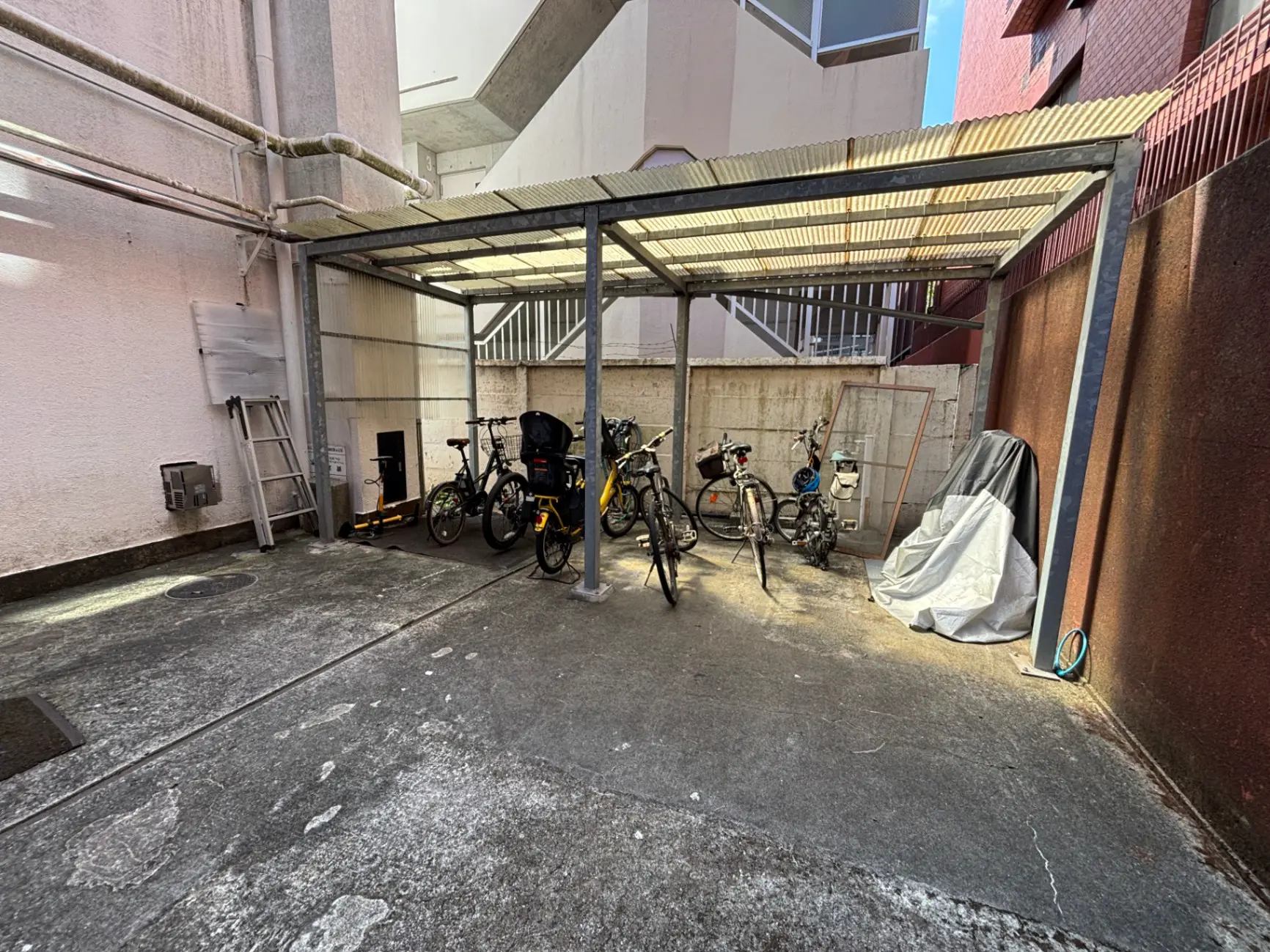 - 自転車が並ぶ屋根付きの駐輪場、左側にははしごが置かれ、右側にはカバーがかかった物体があるアウトドアスペース