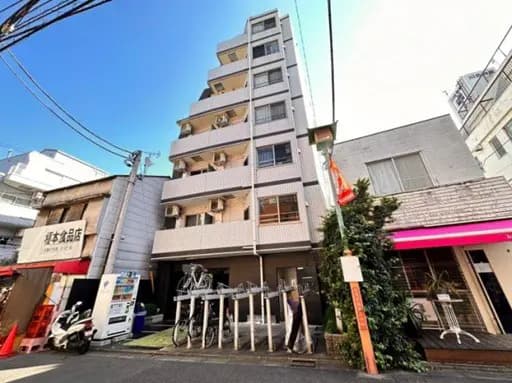 - 都市型の6階建てアパートメントビル、手前には自転車スタンドと周囲の商業施設が並ぶ街並みの風景