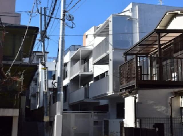 - 都市の一角に建つ現代的なアパートメントブロック、青い空を背景にしたビルの外観と配電線が見える街並み