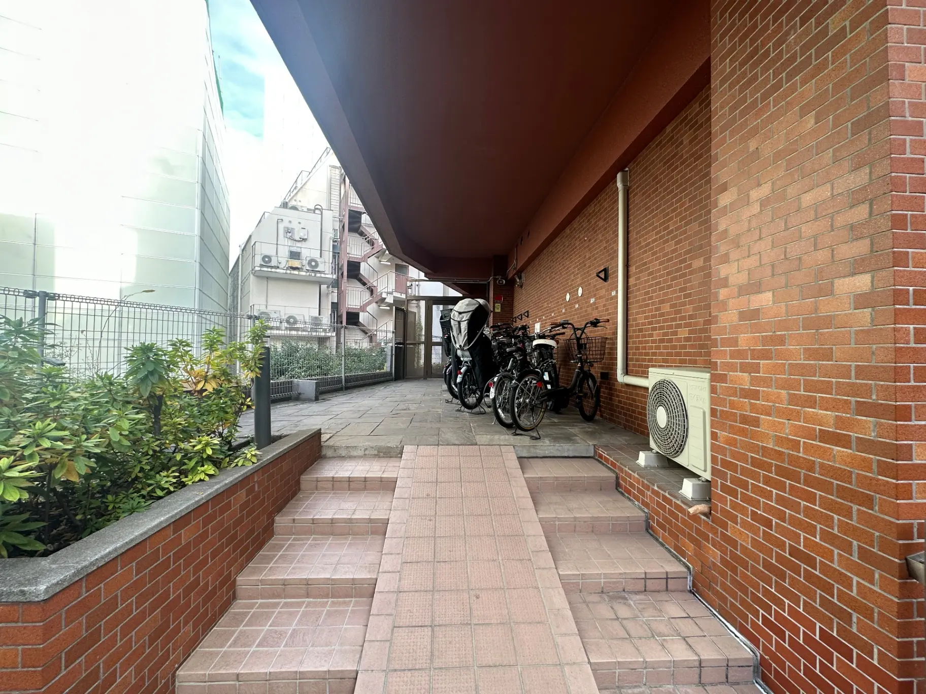 都市の集合住宅のエントランス、レンガ造りの壁、駐輪場に自転車が並ぶ、緑の植栽とタイル張りの階段