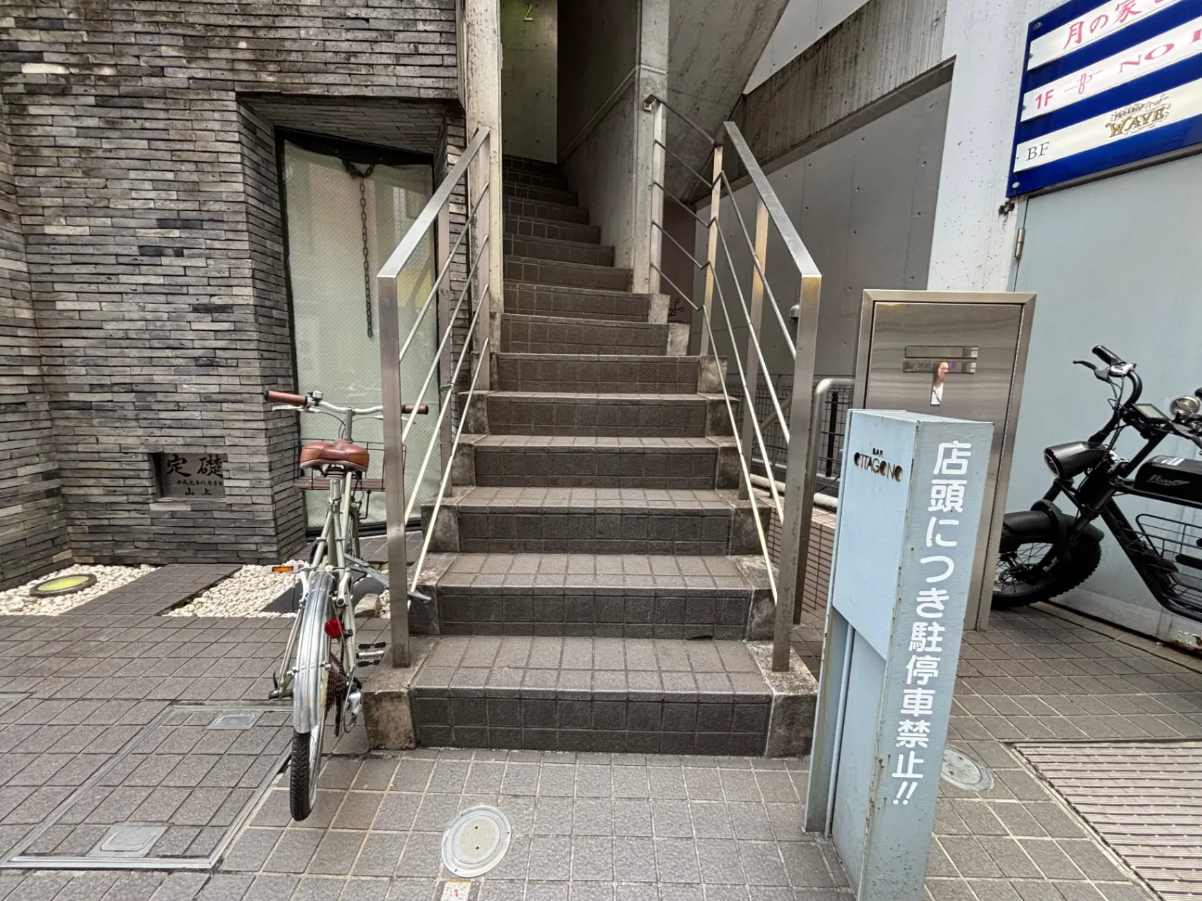 - 複数の自転車と階段がある商業ビルの入り口、ガラスのドアとマンション名が視認できる外観の画像