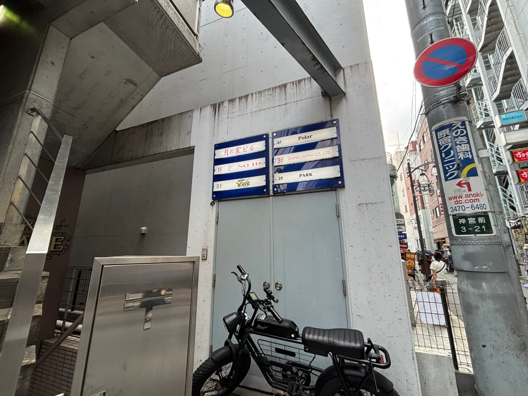- コンクリートの壁と階段、店舗名が掲示された看板、黒い自転車が置かれた雑多な街並みの入口