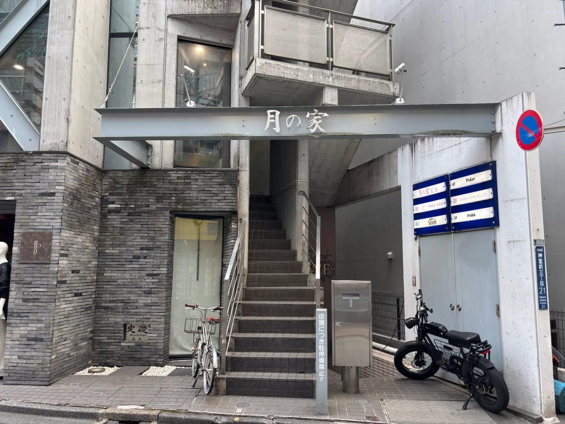 - コンクリート製の外観と階段が特徴的な「月の家」ビルの入り口、周辺には自転車とバイクが停まっている風景