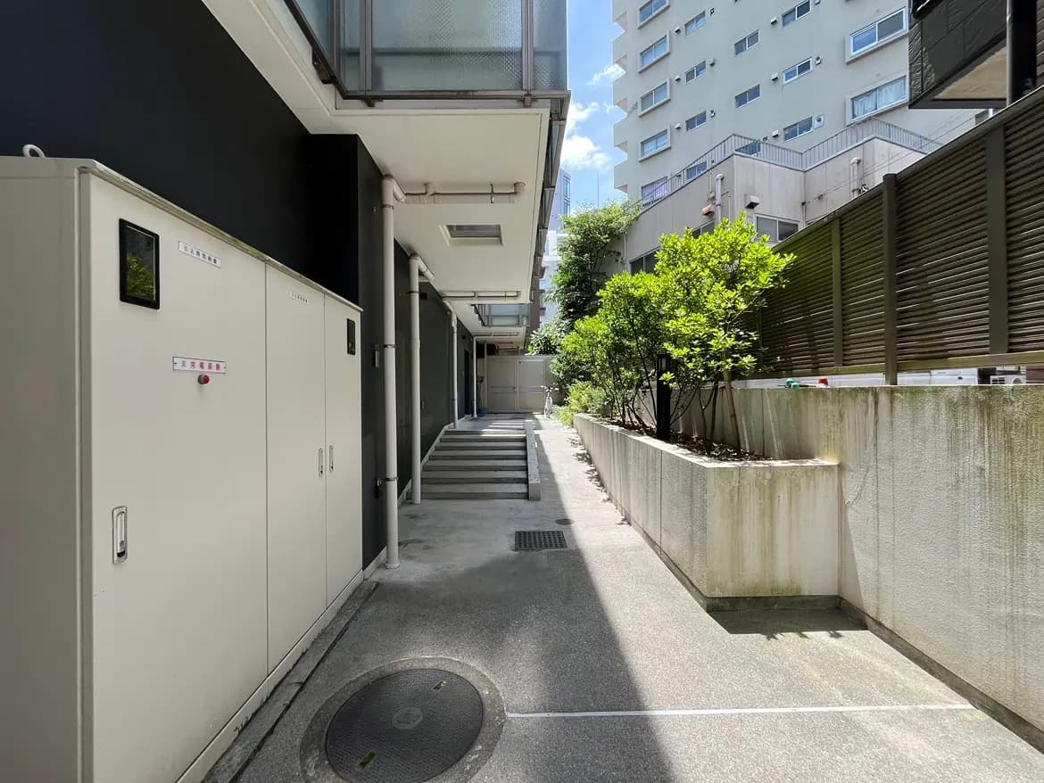 - 日差しが降り注ぐモダンなエントランス、左側に収納ボックスと右側に緑が生い茂る植栽がある通路