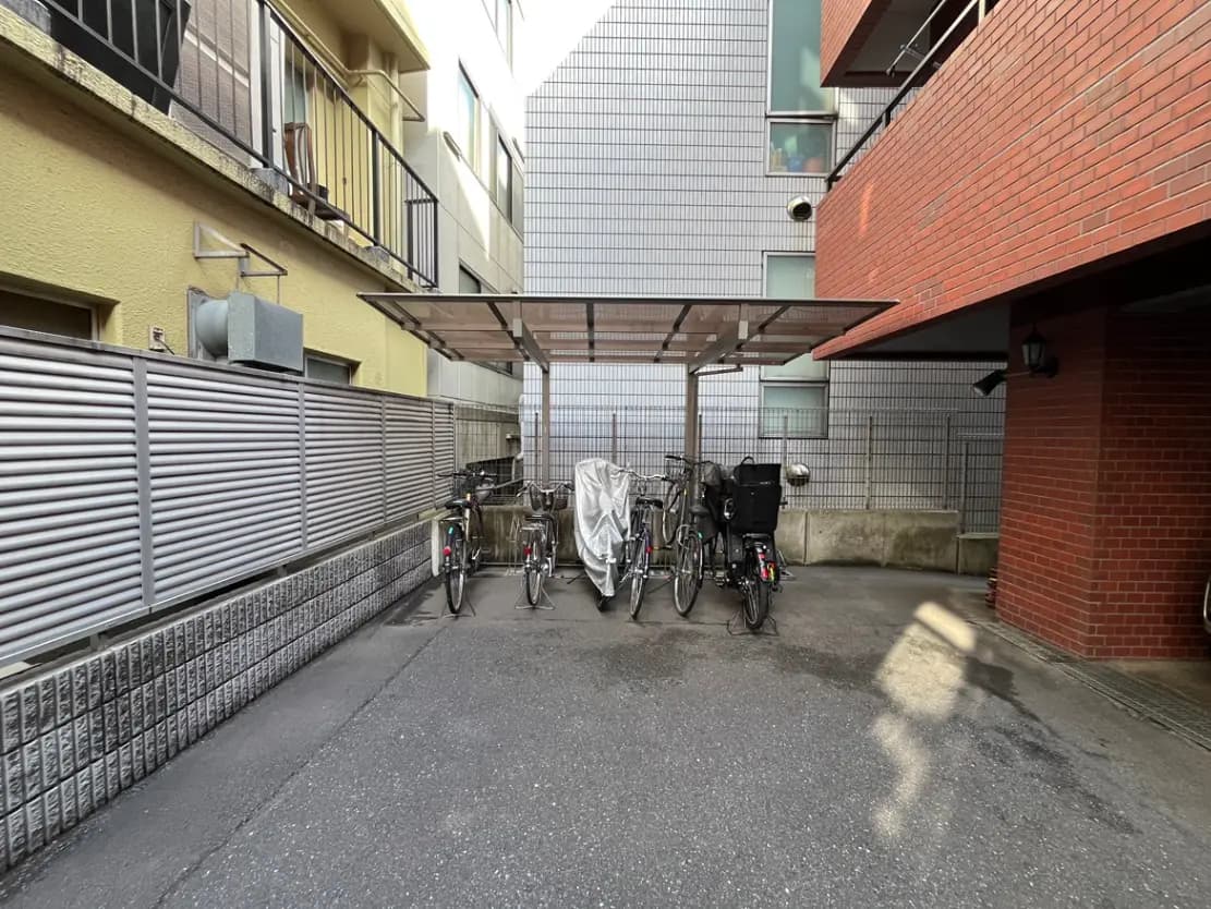 - 自転車置き場の屋根付きスペース、隣接するアパートの外壁が見える、いくつかの自転車が停められた風景