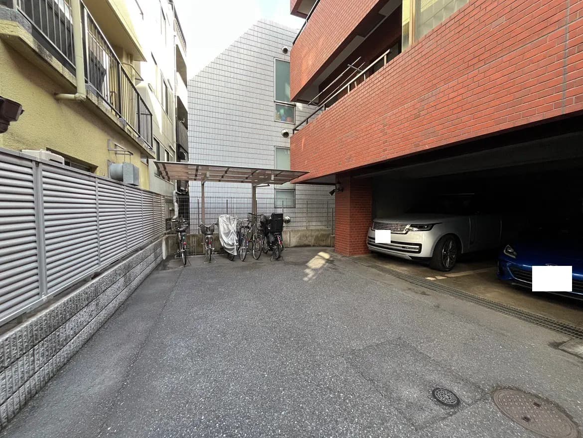 - 駐輪スペースと二台の車両が停まる屋根付き駐車場、周囲にはマンションの外壁とコンクリートの舗装状況が見える静かな環境