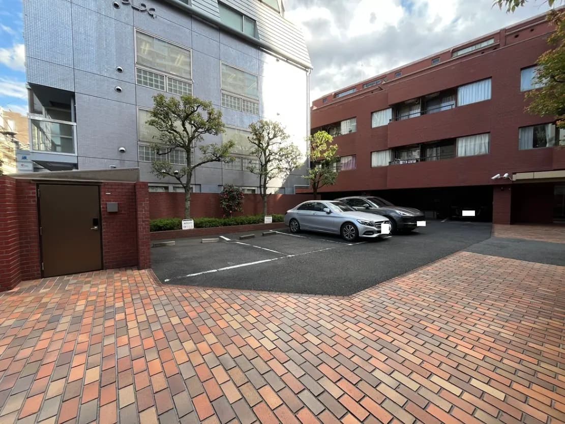 - 都市のマンション前の駐車場、レンガ敷きの路面と隣接する緑の木々、洗練されたエントランスの風景
