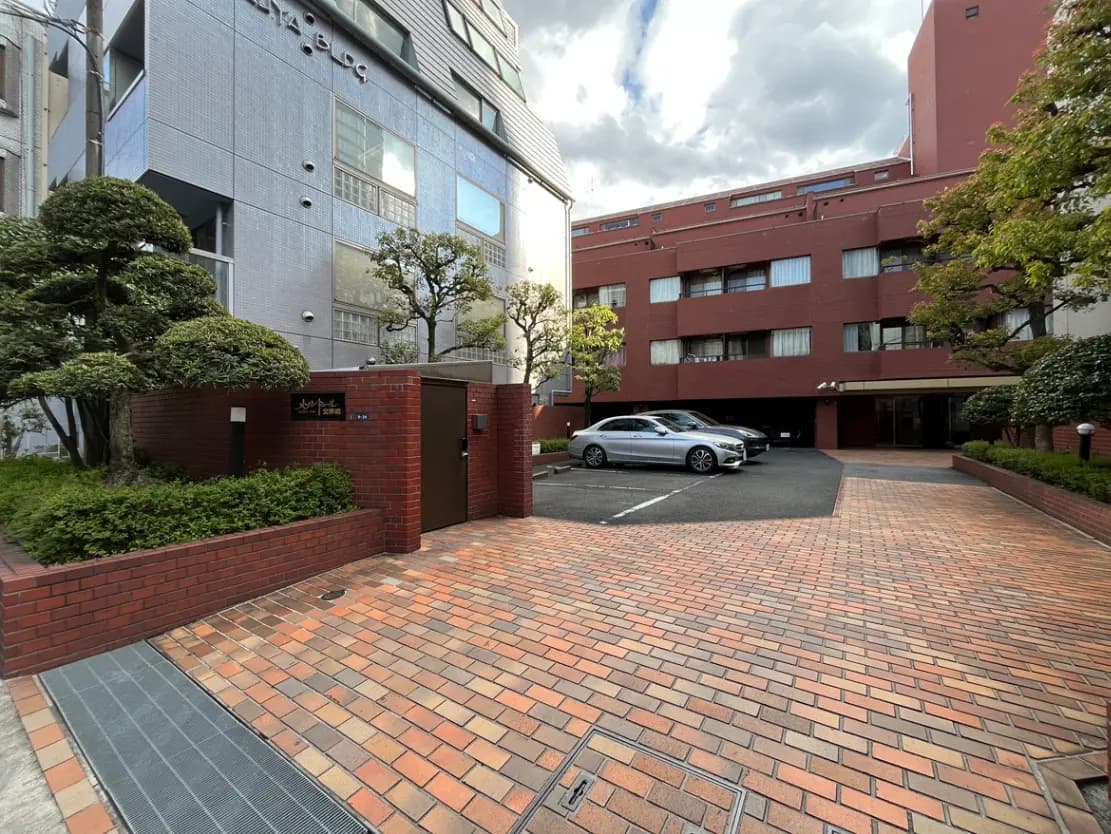 - 都市型マンションのエントランス、煉瓦造りのアプローチと整備された植栽、駐車スペースには車が停まっている景観