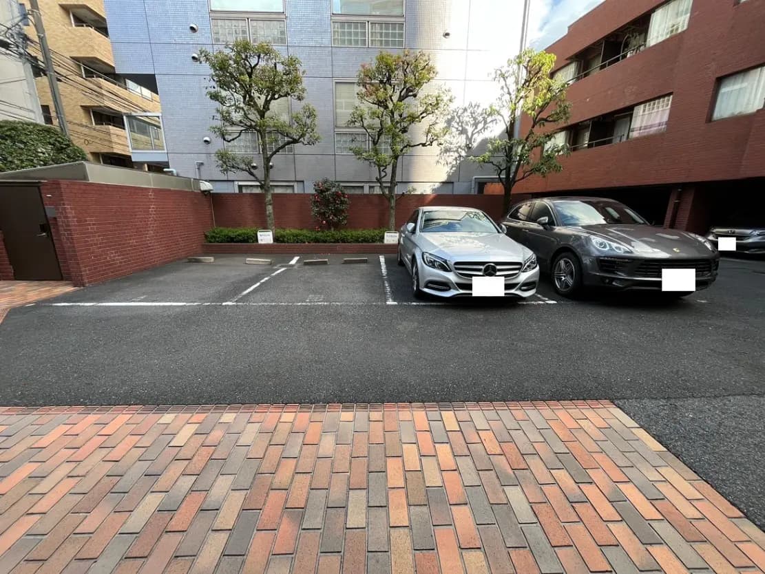 - 駐車場に停車しているシルバーのメルセデス-Benzとグレーのポルシェ、周囲に緑の木とレンガの舗装が施されたエリア