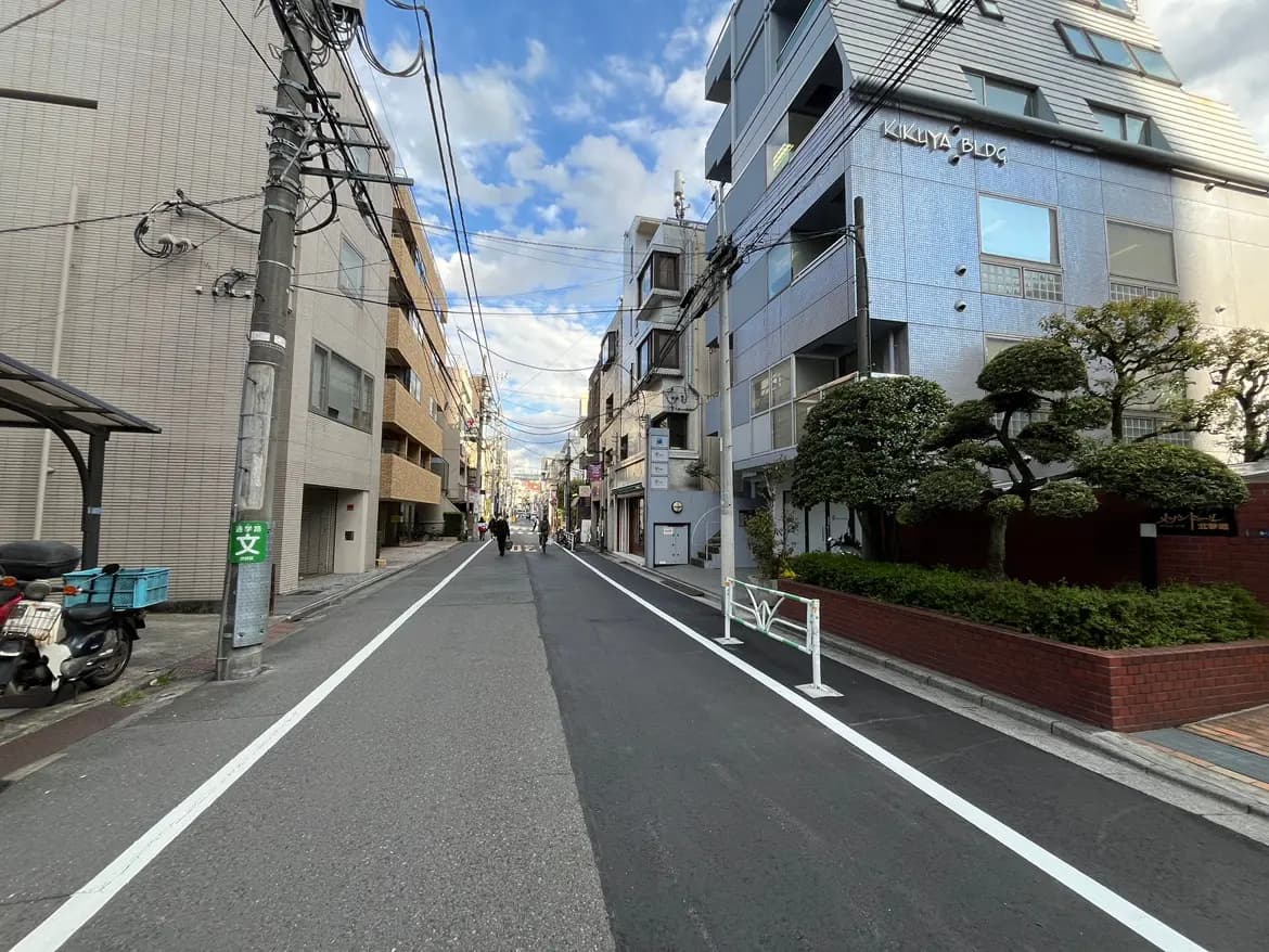 - 都会の一角にある静かな通り、左右に並ぶビルと街路樹、青空に浮かぶ雲が広がる風景