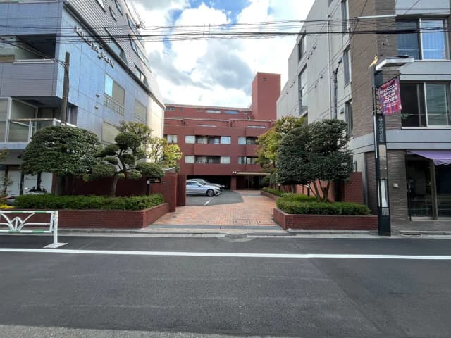 - 都心のマンションエントランス、緑豊かな庭とレンガの花壇、駐車場と青空が広がる風景
