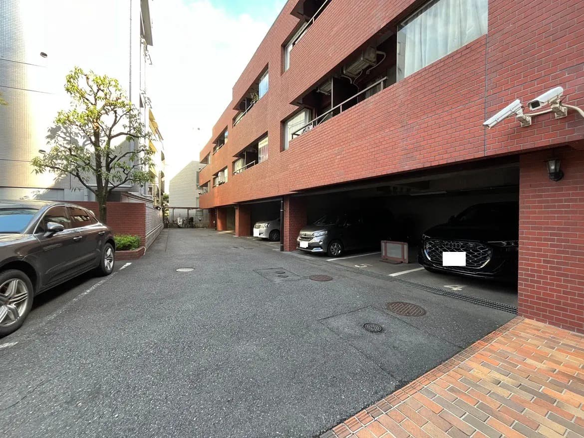 - 駅近の赤レンガの住宅街、車両が停車している駐車場と周囲の緑、周囲に広がる高層ビルの風景