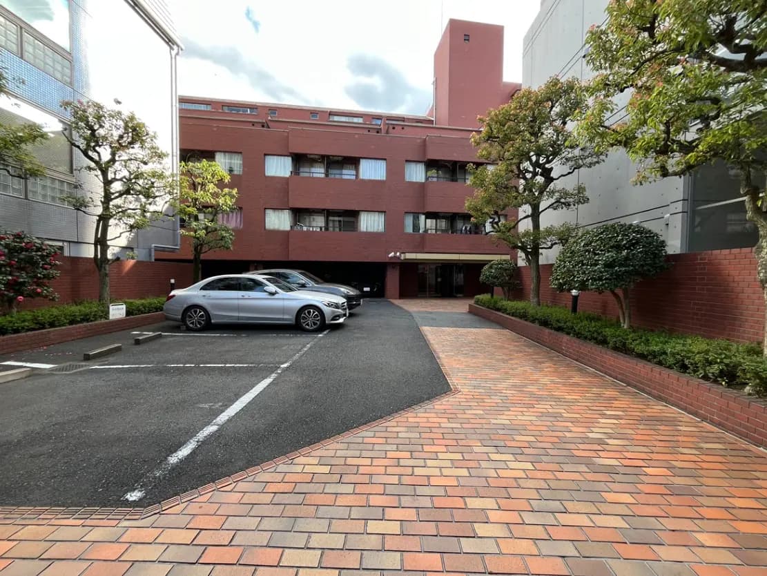 - 駐車場に停まった車と緑の植栽、レンガ舗装の道があるマンションの外観、背景には雲のある空