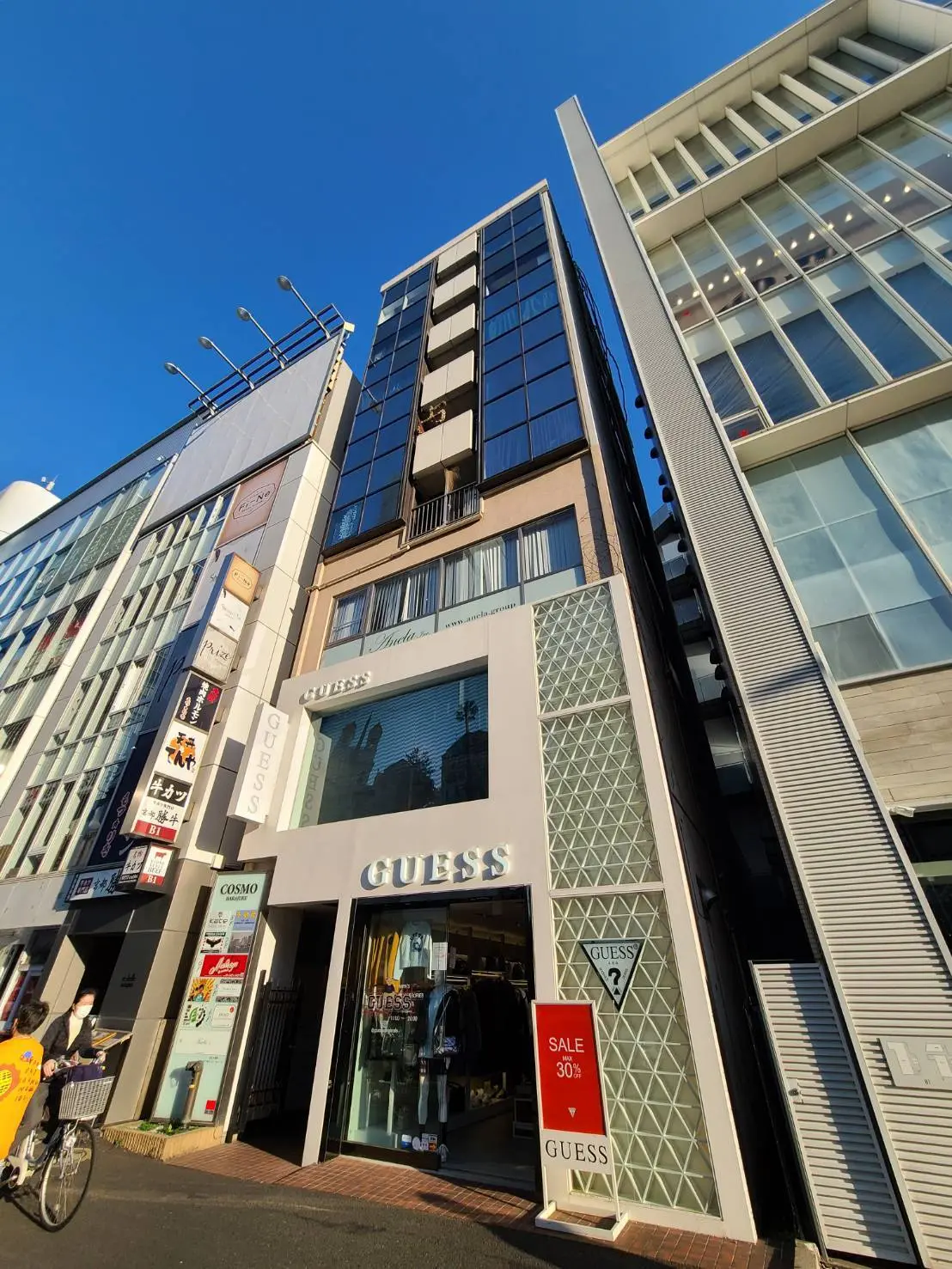 都会のショッピング街に位置するモダンな外観のGUESSブランドの店舗、晴れた日の青空の下、セール告知看板あり
