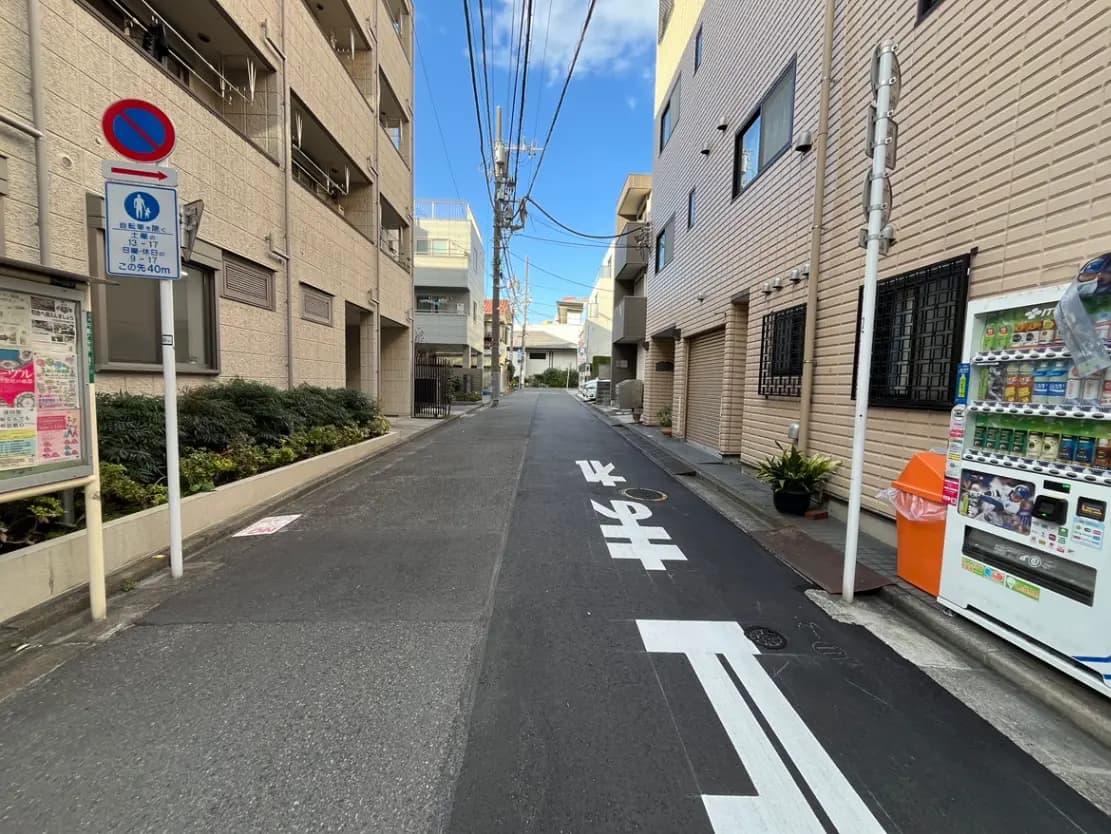 - 都市の住宅街の狭い通り、壁にはアパートメント、街路標識と自動販売機が並ぶ風景