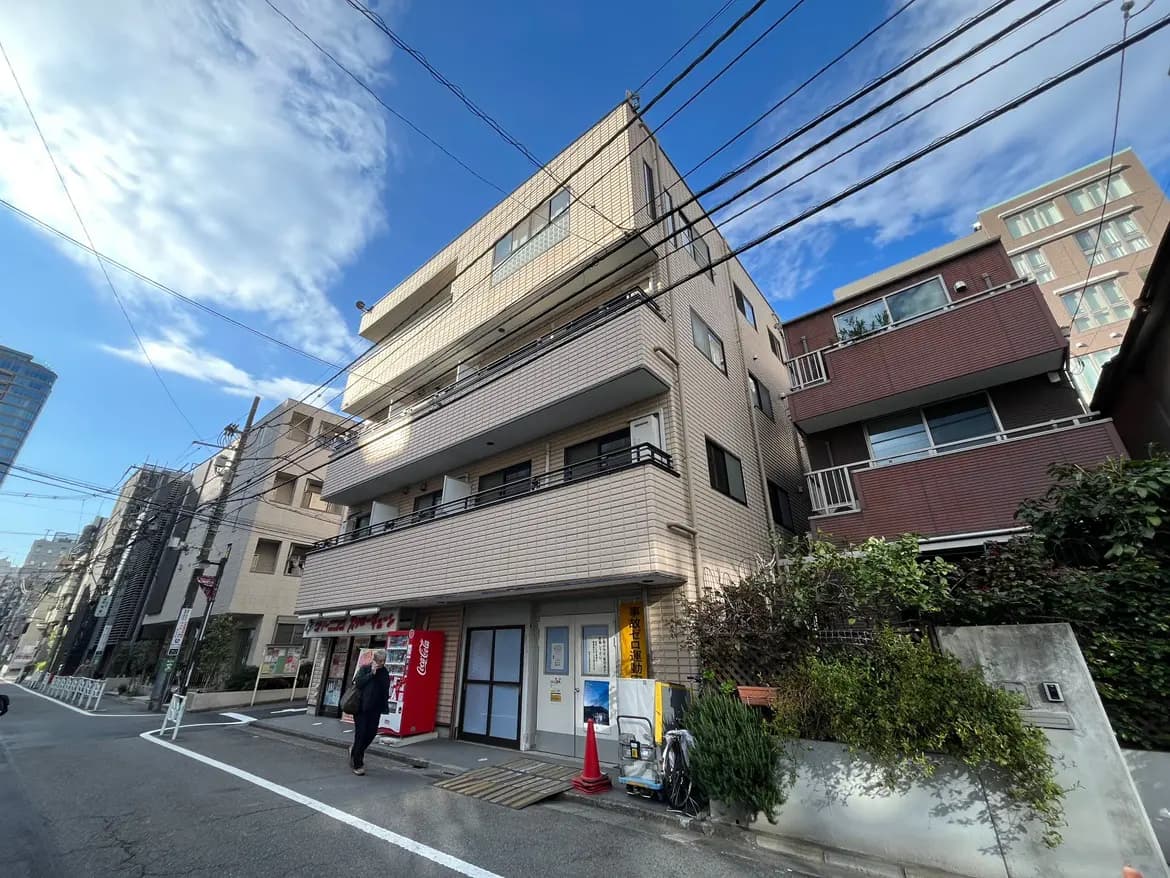 - 都会的な住宅街に位置する3階建てのマンション、周囲には様々な建物が立ち並び、青空と雲が広がる風景