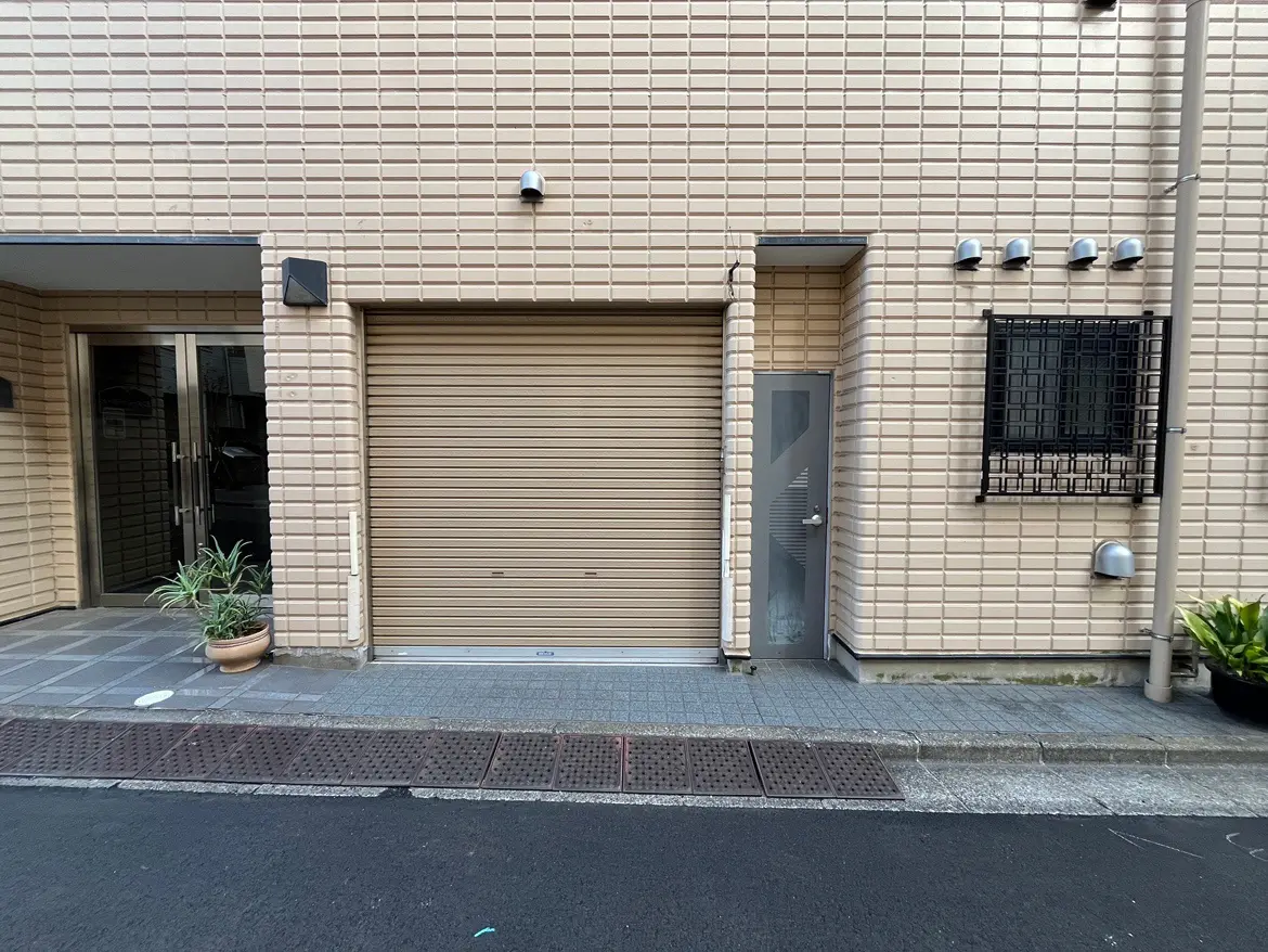 - 明るいベージュの外壁とシャッター付きのガレージ、横に位置するエントランスドア、周囲には植物が配置された現代的な建物の外観