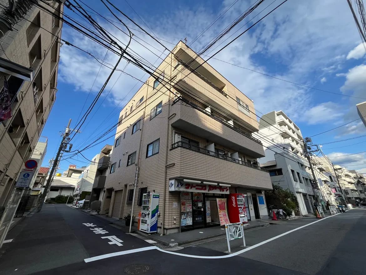 - 多角的な視点から捉えた街の一角、晴れた空の下に立つ多層の建物と電線、通りには自動販売機や看板が並ぶ風景