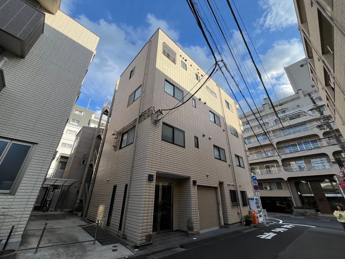 - 都市の住宅街に位置する三階建てのマンション、周囲に他の建物が立ち並び、人々が通行する様子が見える風景