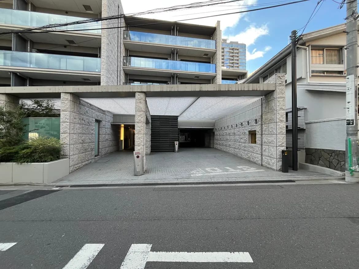 - モダンな建物のエントランス、車が通れるアプローチと石造りの壁、側面には住宅が立ち並ぶ風景