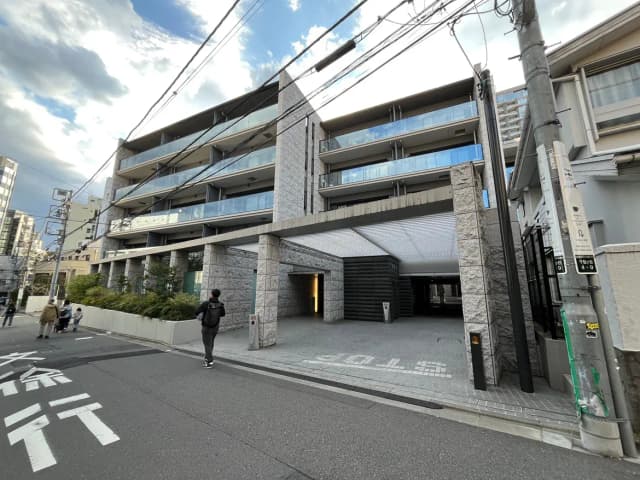 - モダンな外観のマンション、広いアプローチと駐車スペース、周囲に高層ビルが立ち並ぶ街並み