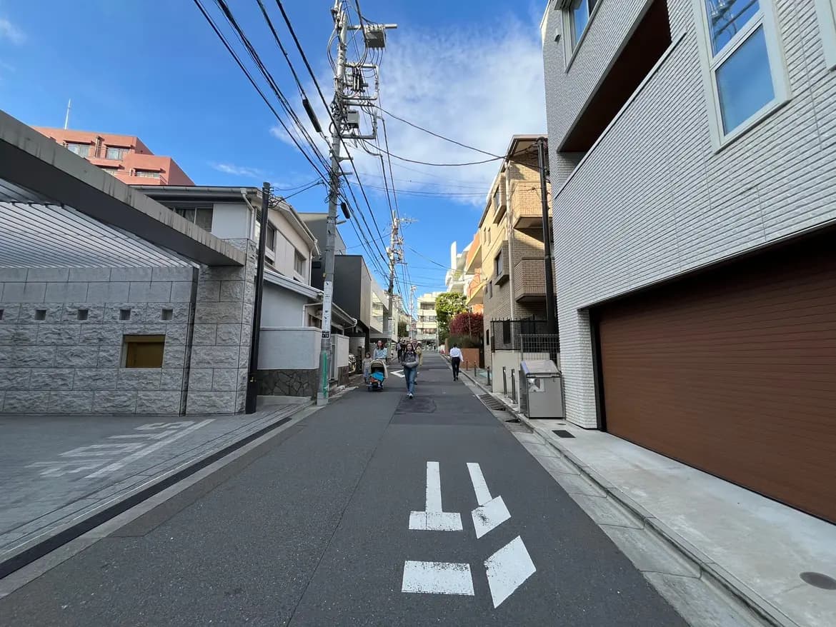 - 都市の住宅街を走る狭い道、周囲には多様な建物と青空が広がる風景、通行人や自転車が見える活気あるシーン