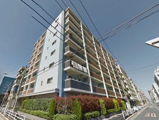 - 大きなマンションの外観、青い外壁とバルコニー、周囲に植栽が整った住宅街の風景