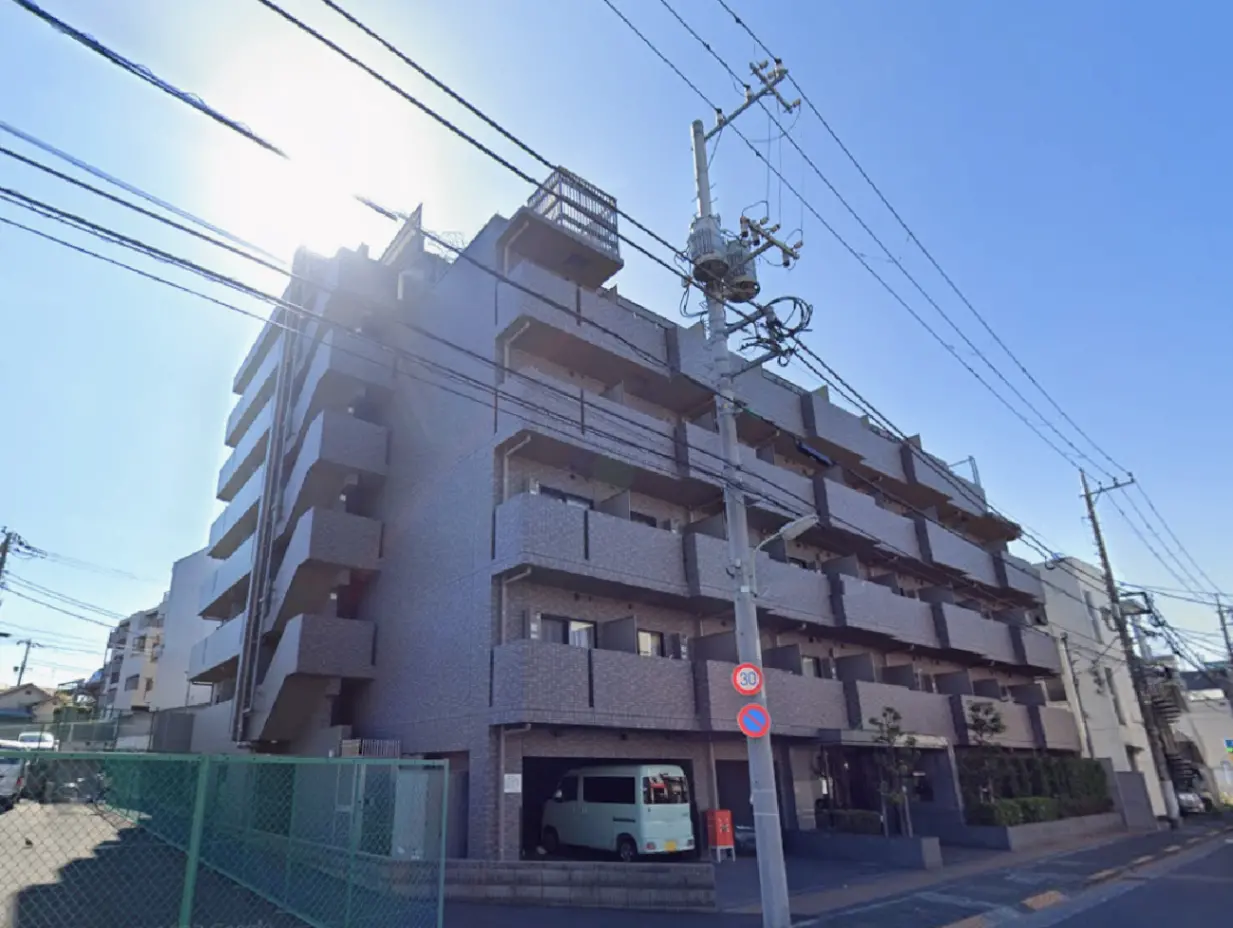 - 青空の下、高層マンションの外観と電柱が並ぶ風景、左側には駐車場とフェンスが見える街並み