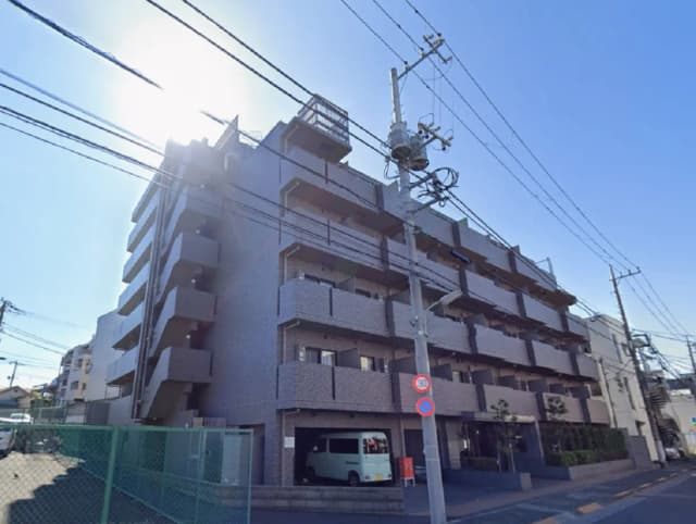 - 青空の下、高層マンションの外観と電柱が並ぶ風景、左側には駐車場とフェンスが見える街並み