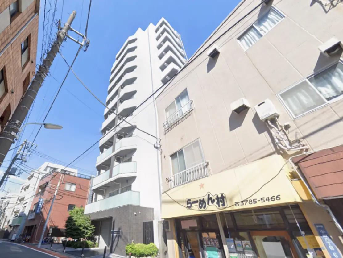 - 明るい青空の下、ラーメン店の看板が目立つ通り沿いのアパートメントビルと周囲の建物、電線が走る街並み