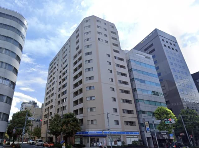 - 都市の高層ビル群の中に立つマンション、周囲には緑の木々や交通信号があり、晴れた空が広がる景色