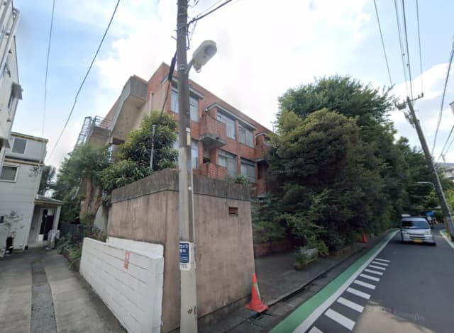 - 緑に囲まれたレンガ造りの建物とその隣の住宅、交通量の少ない道路沿いの風景、周囲の木々が印象的な街並み