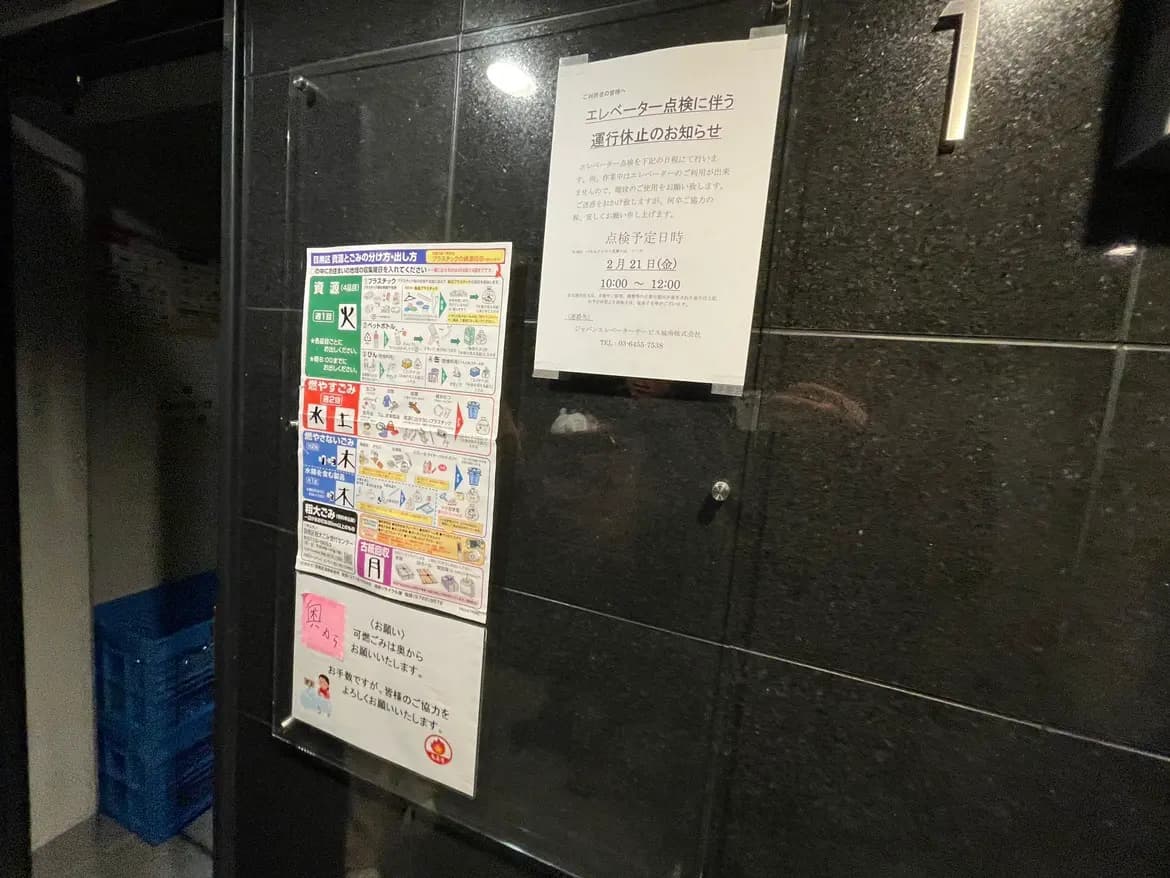 黒い壁のビル入口、注意喚起ポスターや案内掲示、情報ラベルが複数貼られたガラス扉