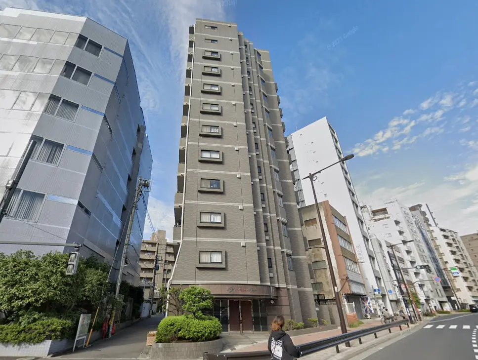 - 都市部に佇む高層マンションの外観、隣接するビルと緑豊かな植え込みがある通りの風景
