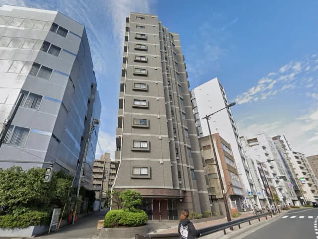 - 都市部に佇む高層マンションの外観、隣接するビルと緑豊かな植え込みがある通りの風景
