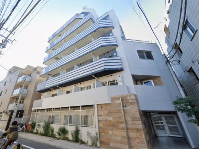 - モダンなデザインのアパートメントビル、白い外壁に青と白のストライプバルコニー、周囲に自転車と緑の植栽が配置された都市の風景