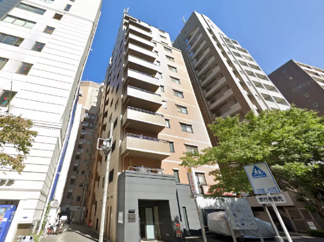 - 都市のマンション外観、隣接する高層ビルと青空、緑の木々がある通りの風景