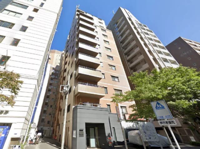 - 都市のマンション外観、隣接する高層ビルと青空、緑の木々がある通りの風景