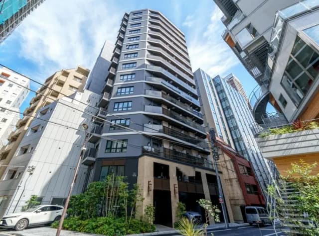 - 都市の中に立つ現代的な高層マンション、周囲のビルと調和した外観、青空と緑の植栽が映える景観