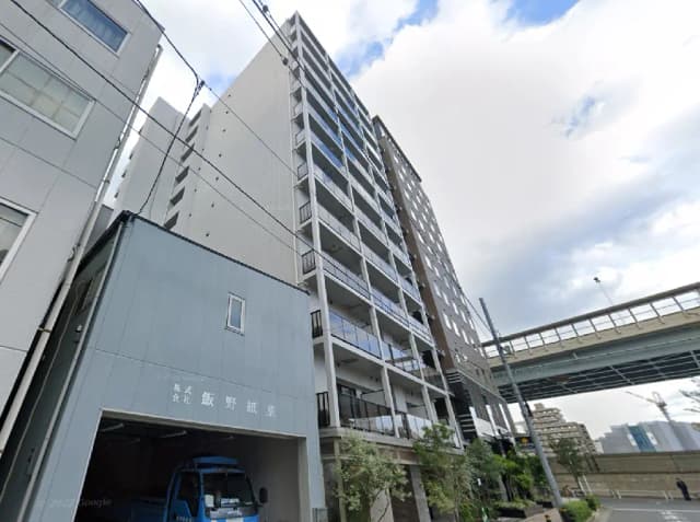 - 高層マンションの外観、周囲にビルが立ち並び、手前には青いトラックと店舗の看板が見える街並み