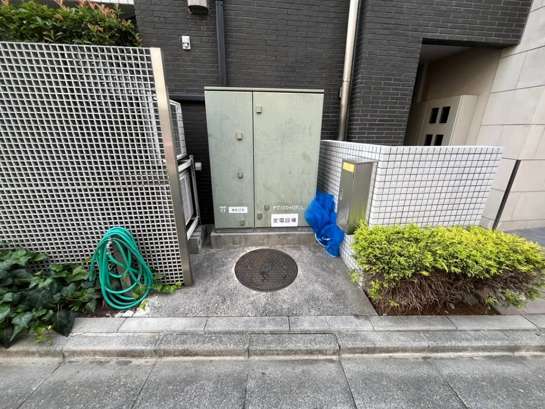 - 駐車場の隅に設置された電気メーターと収納ボックス、周囲には緑の植栽とホースが置かれた景観